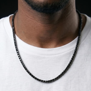 May include: A black tennis chain necklace with small, evenly spaced, black stones. The necklace is worn against a white t-shirt. The chain is a classic jewelry piece.