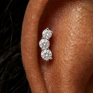 May include: A silver-toned ear piercing featuring three round, clear, faceted stones arranged in a vertical, slightly curved line. The stones are set in a metal base, creating a cluster design that follows the natural contour of the ear cartilage.