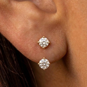 May include: Two round, brilliant-cut clear stones set in gold, worn as a pair of stud earrings. Each stone is secured by four small gold prongs, creating a classic, symmetrical look. The earrings are positioned vertically on the earlobe.