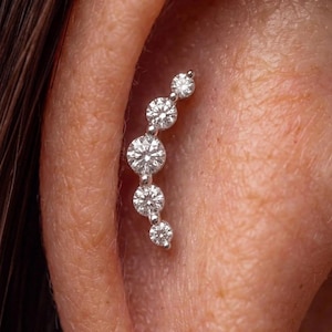 May include: A silver-toned ear climber earring featuring five round, clear, faceted stones arranged in a curved, graduated line. The stones are set in a metallic frame that follows the natural contour of the ear cartilage, creating a sparkling effect.