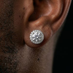 May include: A close-up of a diamond stud earring. The earring features a large, round, clear diamond surrounded by a halo of smaller diamonds. The earring is set in a silver or white gold setting. The earring is in a person's ear.