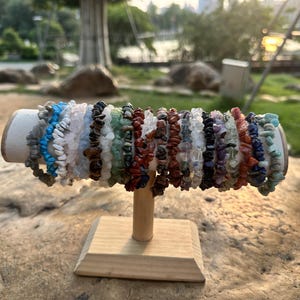 Puede incluir: Exhibición de pulseras de virutas de piedras preciosas en varios colores, incluyendo azul, blanco, verde y rojo. Las pulseras están dispuestas sobre un soporte de madera con una parte superior cilíndrica de color claro y una base cuadrada. El fondo es una escena exterior borrosa.