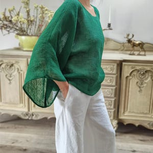 Op de afbeelding: Een persoon draagt een loszittende, smaragdgroene linnen top met wijde mouwen en een witte linnen broek. De top heeft een ronde hals. De broek heeft wijde pijpen. De outfit is casual en comfortabel.
