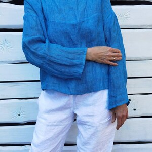 Könnte beinhalten: Ein blaues Leinenoberteil mit langen Ärmeln und rundem Ausschnitt, kombiniert mit einer weißen Leinenhose. Die Kleidung hat eine lockere Passform und eine leicht zerknitterte Textur. Der Hintergrund ist eine weiße Holzwand.