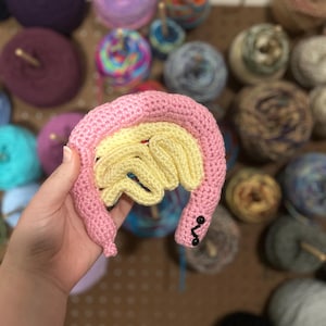 May include: A crocheted anatomical model of a brain, featuring a pink exterior and a yellow interior. The handmade piece includes black button details and is held in front of a backdrop of colorful yarn skeins, suggesting a craft or educational theme.