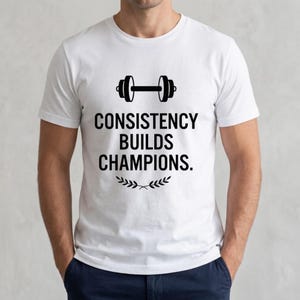 May include: A white short-sleeved t-shirt featuring a black graphic of a dumbbell centered above the text CONSISTENCY BUILDS CHAMPIONS. Below the text is a small black laurel leaf design. The shirt has a crew neckline and a standard fit.
