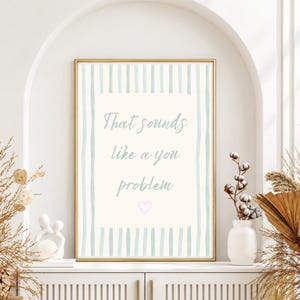 May include: A rectangular wall art print in a gold frame featuring vertical light blue stripes on the border and a cream-colored center. The text reads That sounds like a you problem in light blue script, with a small pink heart icon below.