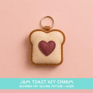 May include: A felt key charm shaped like a slice of toast with a dark pink heart in the center. The toast is light beige with a brown crust border and visible stitching. A gold-toned metal split ring is attached to the top of the charm.