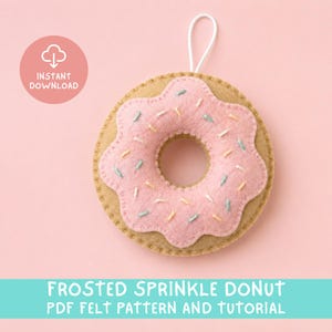 May include: A tan felt donut ornament with pink frosting and small embroidered sprinkles in yellow, blue, and white. A white hanging loop is attached to the top. The image includes a pink circle with the text INSTANT DOWNLOAD and the text FROSTED SPRINKLE DONUT PDF FELT PATTERN AND TUTORIAL.