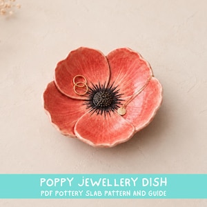 Peut inclure: Un vide-poche en céramique en forme de fleur de coquelicot rouge avec un centre sombre. Le plat contient deux fines bagues dorées et un collier doré délicat avec un petit pendentif circulaire. Le texte POPPY JEWELLERY DISH PDF POTTERY SLAB PATTERN AND GUIDE est visible.