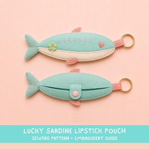 May include: Two felt fish-shaped lipstick pouches in light teal and cream. The top pouch features embroidered details including a green four-leaf clover and a pink heart. The bottom pouch has a pink snap closure and a gold-tone metal keyring.