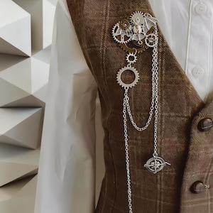 May include: A silver-toned brooch featuring a bat motif, multiple interlocking gears, and dangling chains attached to a small compass charm. The brooch is pinned to a brown plaid vest worn over a white button-down shirt with visible buttons.