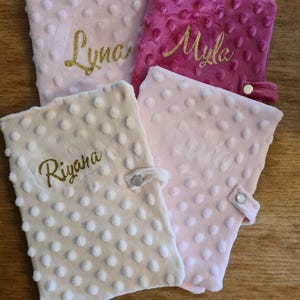 Peut inclure: Quatre couvertures pour bébé en peluche dans les tons de rose et de crème. Chaque couverture a une surface texturée à pois et un nom doré à paillettes : "Lyna", "Myla", "Riyana" et "Olivia". Chaque couverture a une fermeture à bouton-pression.