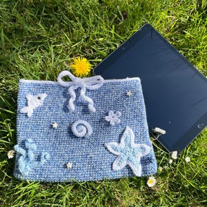 Op de afbeelding: Een blauwe gehaakte tablethoes met witte zeethema versieringen, waaronder een zeester, een vis en een strik. De hoes ligt naast een tablet op groen gras.