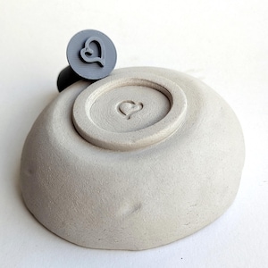 May include: A gray, heart-shaped stamp rests on a light gray ceramic bowl. The stamp's handle is black. The bowl has a heart imprint on the bottom. The stamp is round with a heart design. The bowl is smooth and has a slightly textured surface.