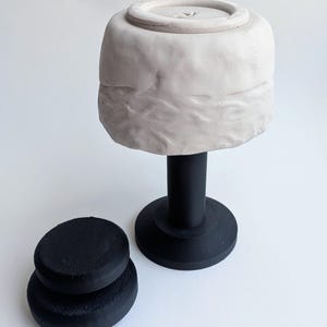 Può includere: Una ciotola in ceramica bianca con una superficie ruvida e irregolare è posizionata capovolta su un supporto cilindrico nero. Accanto al supporto si trova un componente di base circolare nero più piccolo con una finitura opaca e liscia.