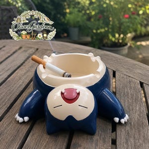 Op de afbeelding: Een donkerblauwe en crèmekleurige keramische asbak in de vorm van een liggend wezen met een wijde open mond, gesloten ogen en witte klauwen. Een brandende sigaret rust in een van de vier inkepingen langs de ronde rand van de asbak.