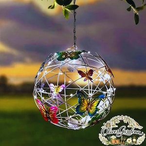 Pode incluir: Um ornamento de jardim esférico de metal com uma estrutura de arame, adornado com detalhes de borboletas coloridas. A esfera é iluminada com pequenas luzes brancas quentes e pende de uma corrente. O fundo apresenta um pôr do sol.