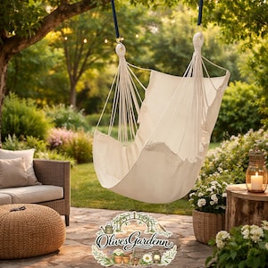 May include: A cream-colored hammock chair hangs from a tree branch in a garden setting. The chair is suspended by white ropes and is surrounded by lush greenery, a wicker sofa, and a stone patio. A candle lantern and a woven basket with flowers are also visible.