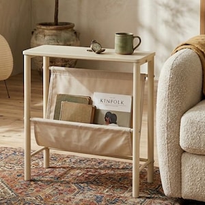 Op de afbeelding: Een crèmekleurige bijzettafel met een ingebouwde tijdschriftenrek. De tafel heeft een groene mok, een kleine klok en een exemplaar van het tijdschrift "Kinfolk". Een plantenpot en een patroonkleed staan op de achtergrond.