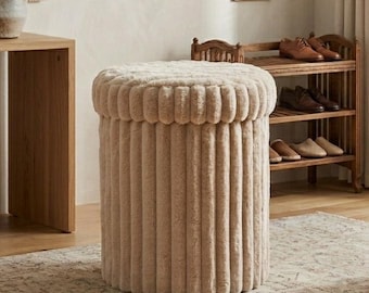 May include: A beige, cylindrical ottoman with a plush, ribbed design. The top and sides have a soft, textured appearance. The ottoman is in a room with framed botanical prints and a wooden shoe rack.