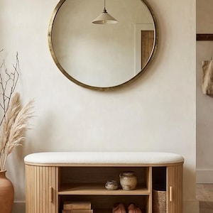 May include: A round gold-framed mirror hangs above a wooden bench with a cream-colored cushion. The bench has slatted doors and open shelves displaying books, shoes, and decorative items. A vase with dried plants and a woven basket add to the decor.