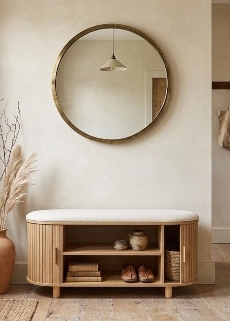 May include: A round gold-framed mirror hangs above a wooden bench with a cream-colored cushion. The bench has slatted doors and open shelves displaying books, shoes, and decorative items. A vase with dried plants and a woven basket add to the decor.