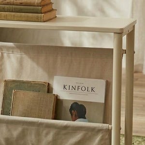 May include: Off-white magazine rack with a top shelf holding three vintage books. The rack has a fabric pocket containing a "KINFOLK" magazine and two other books. The rack is made of metal.