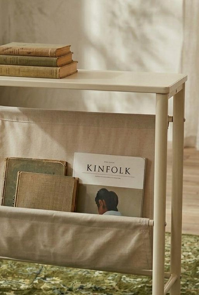 May include: Off-white magazine rack with a top shelf holding three vintage books. The rack has a fabric pocket containing a "KINFOLK" magazine and two other books. The rack is made of metal.