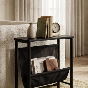 May include: A black side table with a built-in magazine rack. The table holds a small vase, several books, and a clock. The magazine rack is filled with magazines and a leather wallet. The table is set on a patterned rug.
