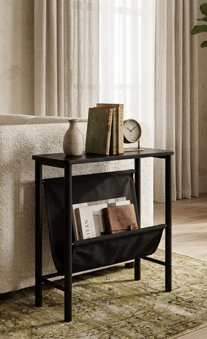 May include: A black side table with a built-in magazine rack. The table holds a small vase, several books, and a clock. The magazine rack is filled with magazines and a leather wallet. The table is set on a patterned rug.