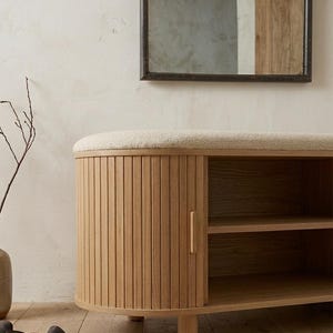 May include: A light-colored wooden storage bench with a cushioned top and a slatted door. The bench has two shelves inside and is set against a neutral-toned wall. A mirror hangs above the bench.