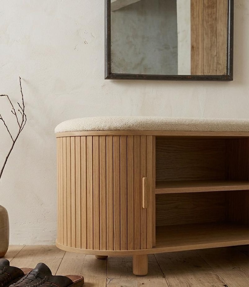 May include: A light-colored wooden storage bench with a cushioned top and a slatted door. The bench has two shelves inside and is set against a neutral-toned wall. A mirror hangs above the bench.