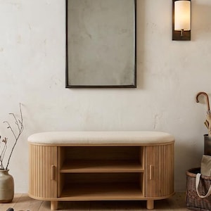 May include: A rectangular mirror with a dark frame hangs above a wooden bench with a cream-colored cushioned top. The bench has two doors and open shelves. A wall sconce and a vase with branches are also visible.
