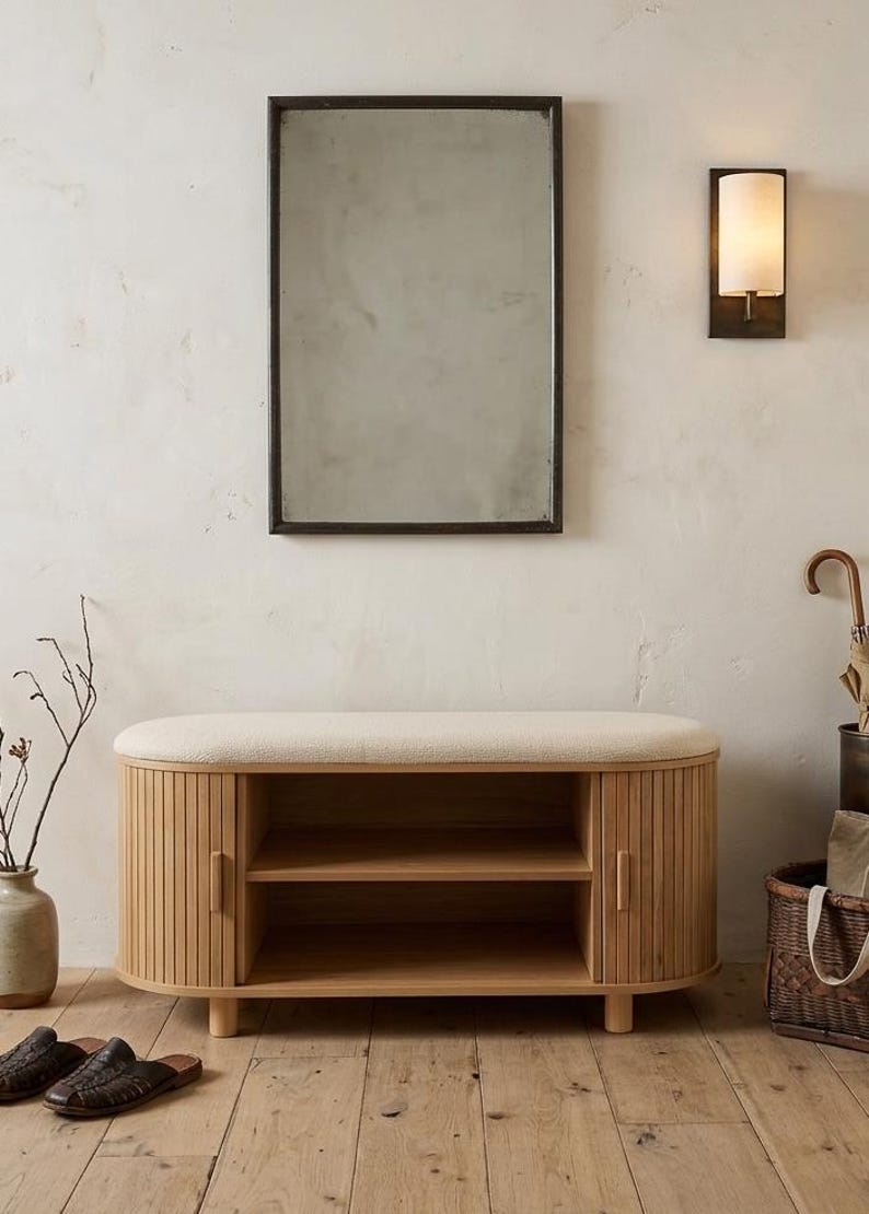 May include: A rectangular mirror with a dark frame hangs above a wooden bench with a cream-colored cushioned top. The bench has two doors and open shelves. A wall sconce and a vase with branches are also visible.