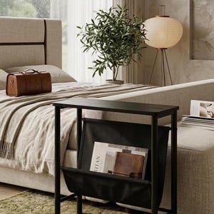 May include: A black side table with a magazine rack, holding books and a leather-bound journal. The table is in a bedroom setting, next to a bed with a brown leather suitcase. A green rug is on the floor.