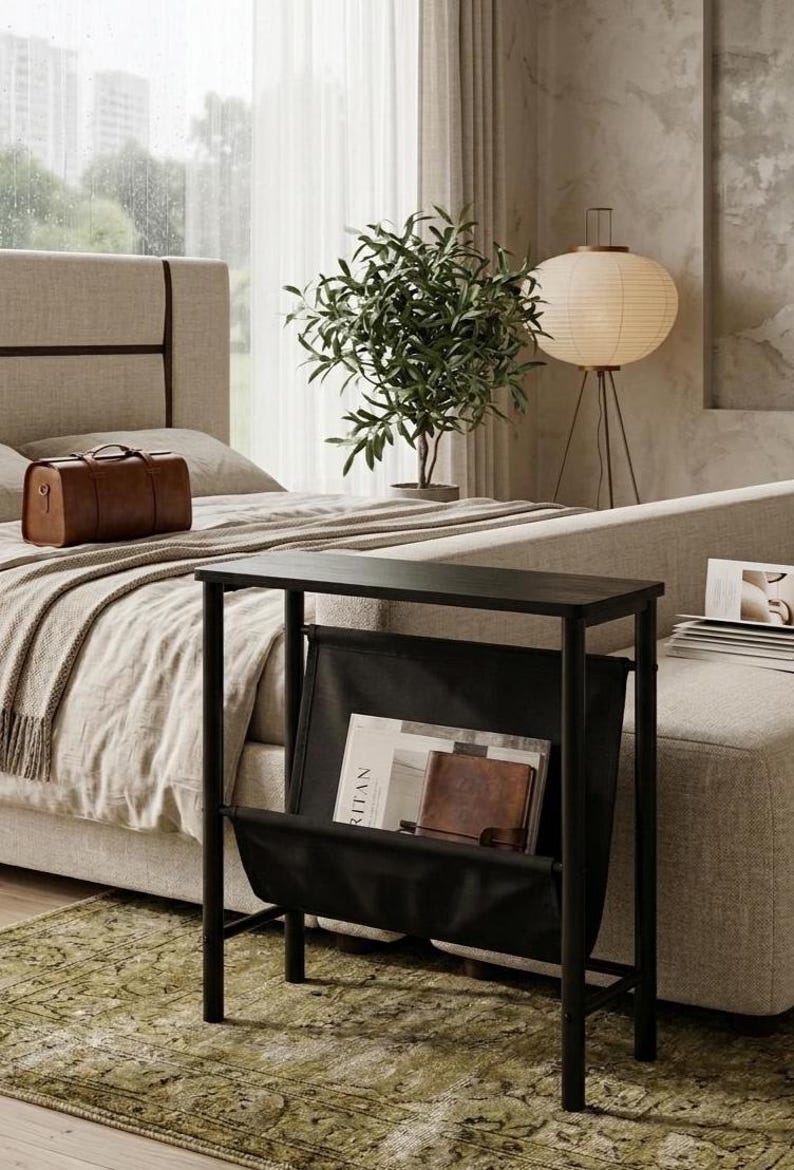 May include: A black side table with a magazine rack, holding books and a leather-bound journal. The table is in a bedroom setting, next to a bed with a brown leather suitcase. A green rug is on the floor.
