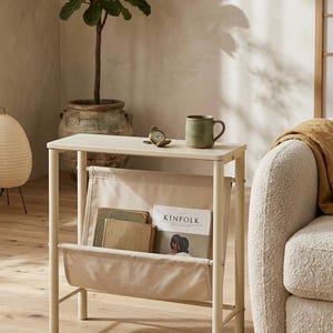 May include: A cream-colored side table with a built-in magazine rack. The table holds a green mug, a small clock, and a copy of "Kinfolk" magazine. A potted plant and a beige sofa are in the background, creating a cozy, neutral-toned living space.