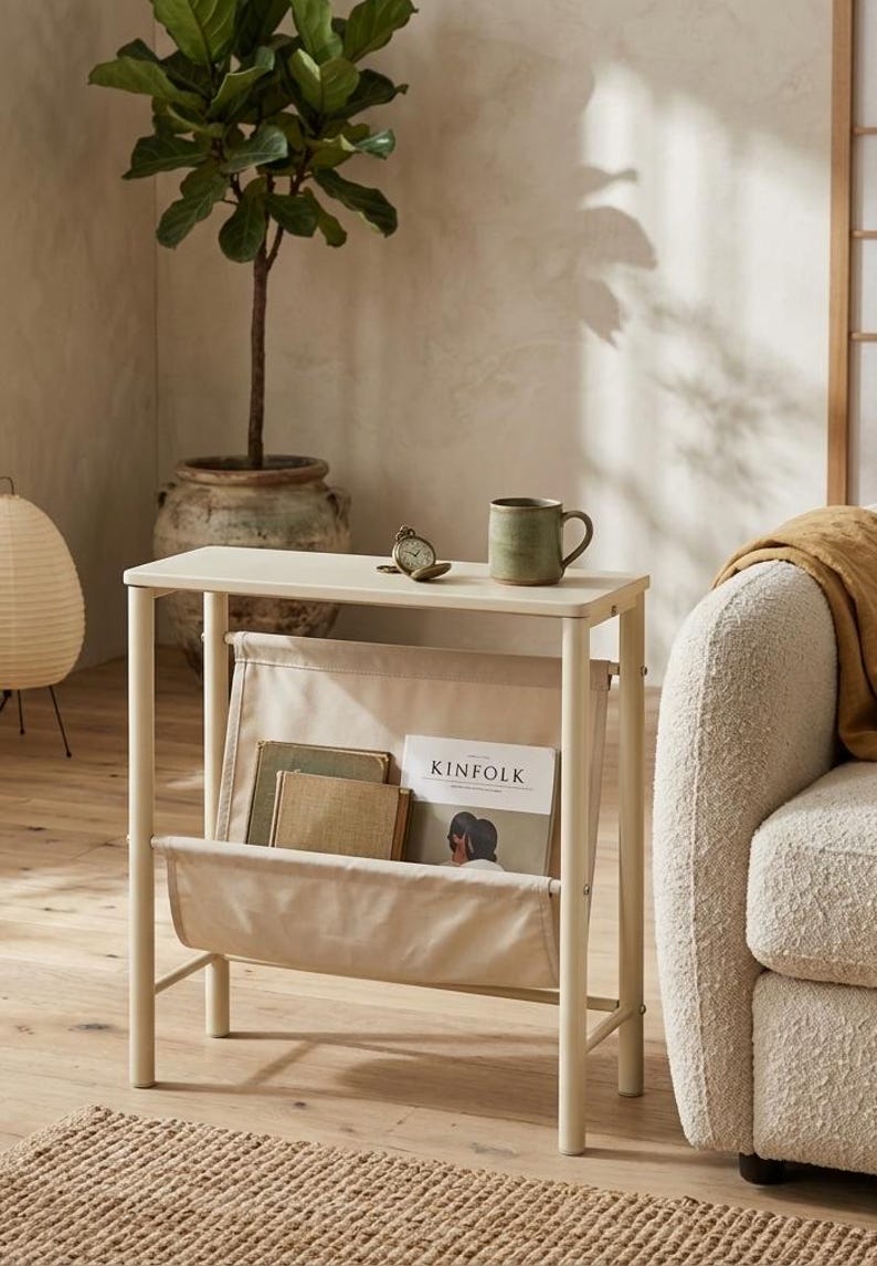 May include: A cream-colored side table with a built-in magazine rack. The table holds a green mug, a small clock, and a copy of "Kinfolk" magazine. A potted plant and a beige sofa are in the background, creating a cozy, neutral-toned living space.