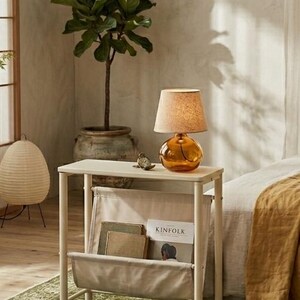 May include: A small, cream-colored side table with a built-in magazine rack. A round, amber-colored lamp sits on top, next to a small decorative bird. A potted plant and a green rug are in the background. The magazine rack holds books and a copy of "Kinfolk".