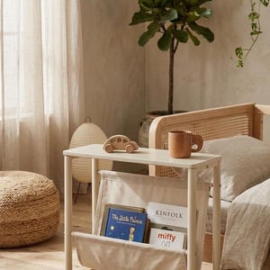 May include: A small, off-white side table with a canvas magazine rack. A wooden toy car and a brown mug sit on top. Books include "The Little Prince" and "Kinfolk". The scene is in a neutral-toned bedroom with a plant.