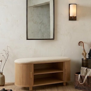 May include: A wooden shoe bench with a cushioned top and slatted doors, paired with a rectangular mirror in a dark frame. A wall sconce and a woven basket complete the entryway decor. The bench has two shelves for storage.