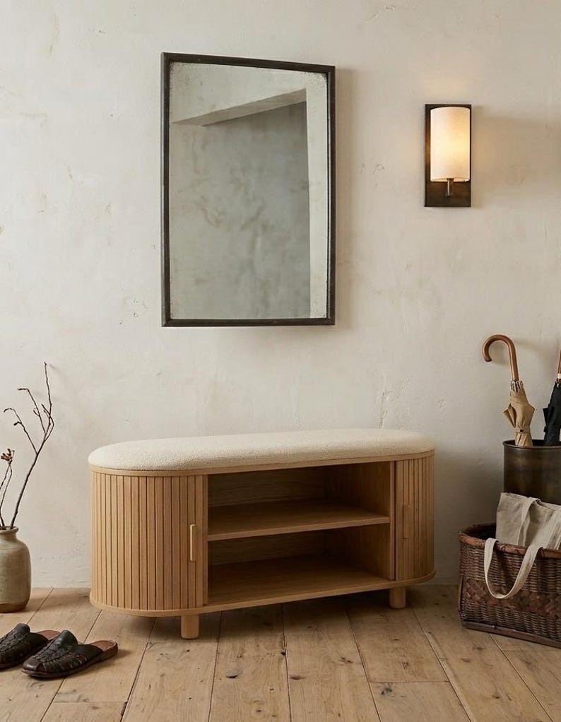 May include: A wooden shoe bench with a cushioned top and slatted doors, paired with a rectangular mirror in a dark frame. A wall sconce and a woven basket complete the entryway decor. The bench has two shelves for storage.