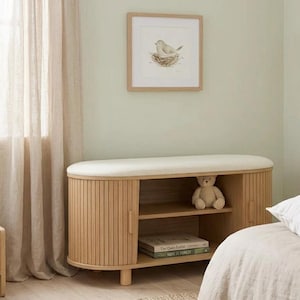 May include: A light wood bench with a cream-colored cushioned top and fluted sides. The bench has two doors and an open shelf. A framed bird print hangs on the wall above the bench. A teddy bear sits inside the bench.