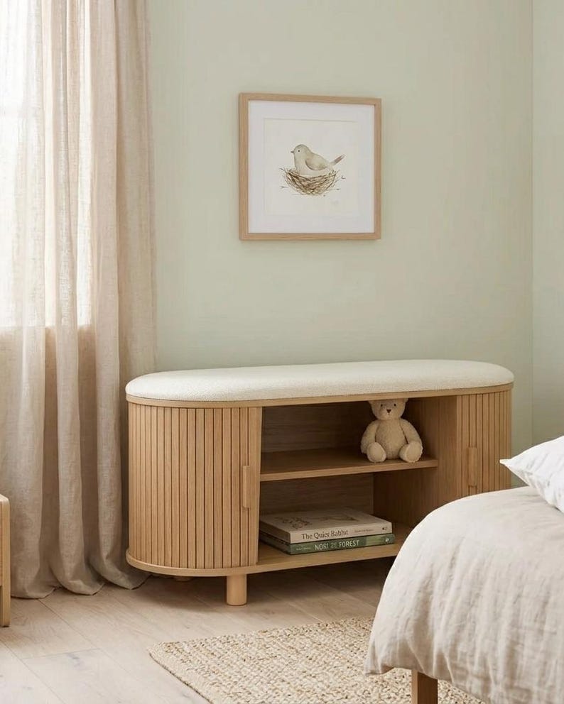 May include: A light wood bench with a cream-colored cushioned top and fluted sides. The bench has two doors and an open shelf. A framed bird print hangs on the wall above the bench. A teddy bear sits inside the bench.