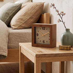 Peut inclure: Un réveil carré en bois avec un cadran beige et des aiguilles dorées est posé sur une table de chevet en bois. L'horloge affiche les chiffres 12, 3, 6 et 9. Un petit vase vert avec des fleurs et une boîte dorée sont également sur la table.