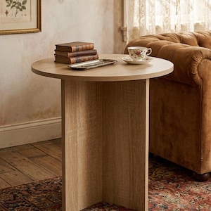 Op de afbeelding: Een ronde, lichtgekleurde houten bijzettafel met een voetstuk. Op de tafel liggen boeken, een zilveren dienblad en een bloemen theekopje en schotel. De tafel staat in een kamer met een bruine bank en een patroonkleed.