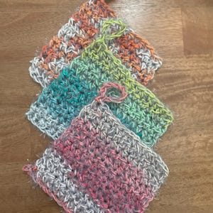 Puede incluir: Cuatro paños de cocina de ganchillo hechos a mano en tonos naranja, verde, turquesa y rosa. Cada paño de cocina cuadrado tiene un lazo para colgar. Los paños de cocina están hechos con un hilo texturizado y están colocados sobre una superficie de madera.