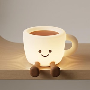Puede incluir: Una lámpara brillante con forma de taza de color blanco roto, con un asa redondeada y lisa y un rostro impreso sencillo con dos pequeños ojos negros y una sonrisa curva. Dos piernas cilíndricas marrones sobresalen de la base.