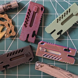 May include: Four rectangular utility tools with sliding mechanisms, featuring a textured finish in metallic purple, matte olive green, matte black, and iridescent rainbow colours, alongside a gold bicycle chain keyring and two silver multi-tools.